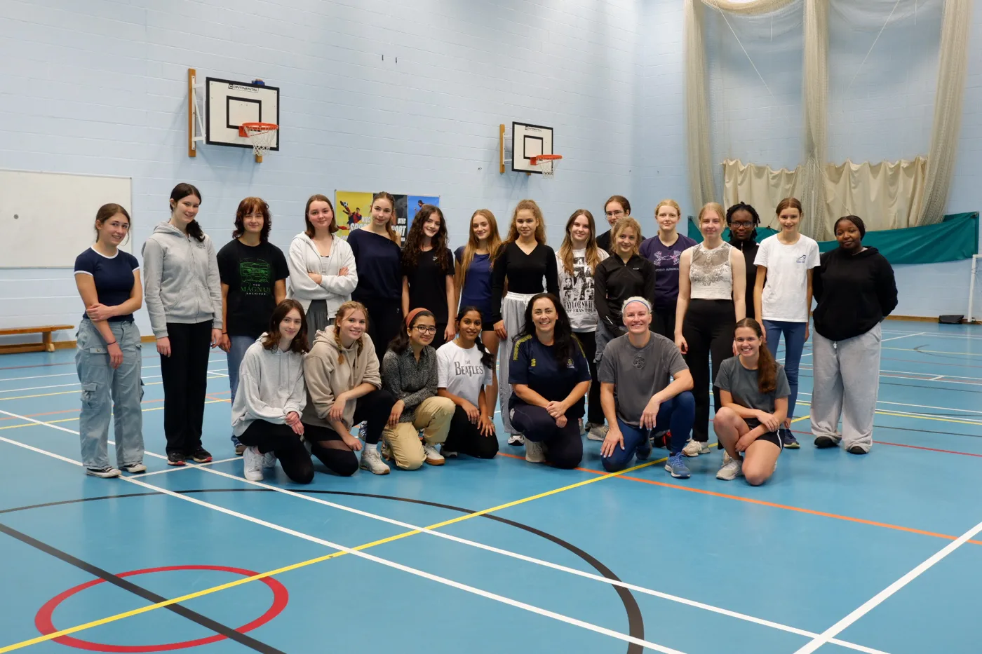 A group of about 25 girls and women standing and kneeling in rows in a sports hall with basketball hoops